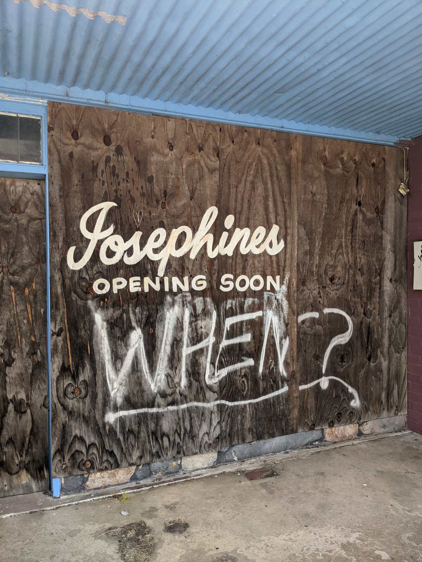 A boarded-up shopfront painted 'opening soon,' with graffiti asking 'when?'